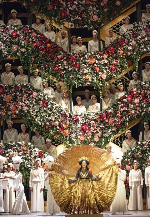 Eine Person in einem goldenen, sonnenähnlichen Kostüm im Vordergrund. Dahinter eine Blumenkulisse mit Darstellern in weißen Gewändern, die auf mehreren Ebenen angeordnet und von leuchtenden Blumen und Grünpflanzen umgeben sind.