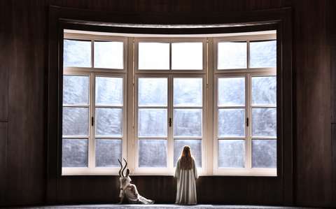 Eine Person in einem weißen Gewand steht vor einem großen, rechteckigen Fenster mit mehreren Scheiben und blickt nach draußen. Eine kleine, gehörnte Statue oder Figur steht neben ihr auf dem Boden.
