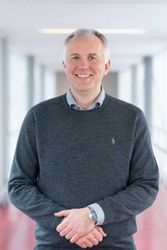 Prof. Dr. Gregor Szycik in grauem Pullover in einem Raum mit Fenstern.