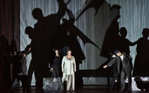 Darsteller auf der Bühne werfen bei dramatischer Beleuchtung Schatten auf einen großen Vorhanghintergrund. Die Szene zeigt in der Mitte eine Frau in einem langen Kleid, umgeben von Figuren in Theaterposen, die eine dynamische und stimmungsvolle Atmosphäre schaffen.