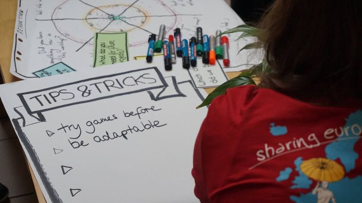 Plakat mit der Überschrift "Tips and Tricks". Davor sitzt eine Frau im EuroPeer-T-Shirt mit der Aufschrift "Sharing Europe"