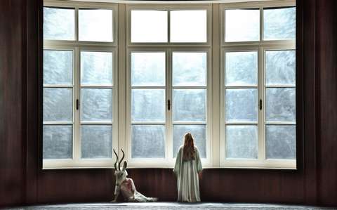 Eine Person in einem langen Kleid steht und blickt aus einem großen, gewölbten Fenster mit Milchglasscheiben. Neben ihr sitzt eine Gestalt mit spitzer Maske und Robe auf der Fensterbank. Sonnenlicht fällt durch das Fenster und wirft einen sanften Schein hinein.