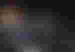 Eine Gruppe von Darstellern, alle in schwarzen Mänteln und Zylindern, umringt eine Person in der Mitte, die mit ausdrucksstarkem Gesicht nach oben blickt und möglicherweise singt. Der Hintergrund ist dunkel, mit einer golden beleuchteten Box auf der linken Seite.