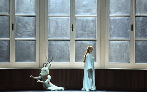 Eine Theaterszene mit einer Person in einem weißen Gewand, die in der Nähe großer Fenster steht, und einer anderen Person mit einem weißen Hirschkopf, die auf der Bühne kniet. Die Beleuchtung ist sanft und schafft eine surreale Atmosphäre.