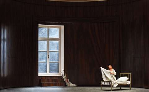 Ein minimalistisches Theaterset mit einer großen, dunklen Holzkulisse. Ein Fenster auf der linken Seite sorgt für Licht. Eine Frau sitzt auf einem einfachen Bett und hält ein weißes Laken in der Hand. Ein weißes Kleid und ein Geweih liegen auf dem Boden neben dem Fenster.