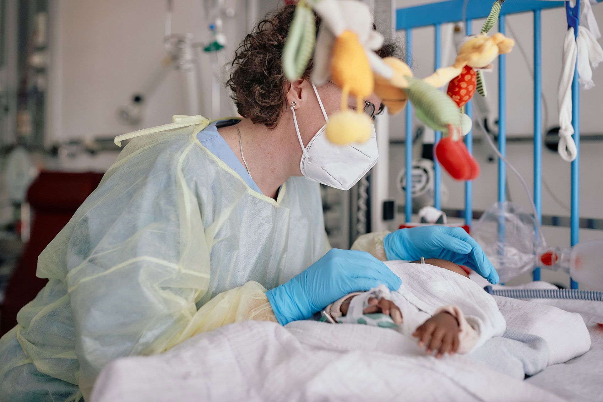 Fotos eines Kindes in einem Krankenhausbett. Eine Krankenhausmitarbeiterin in steriler Kleidung beugt sich über das Kind.
